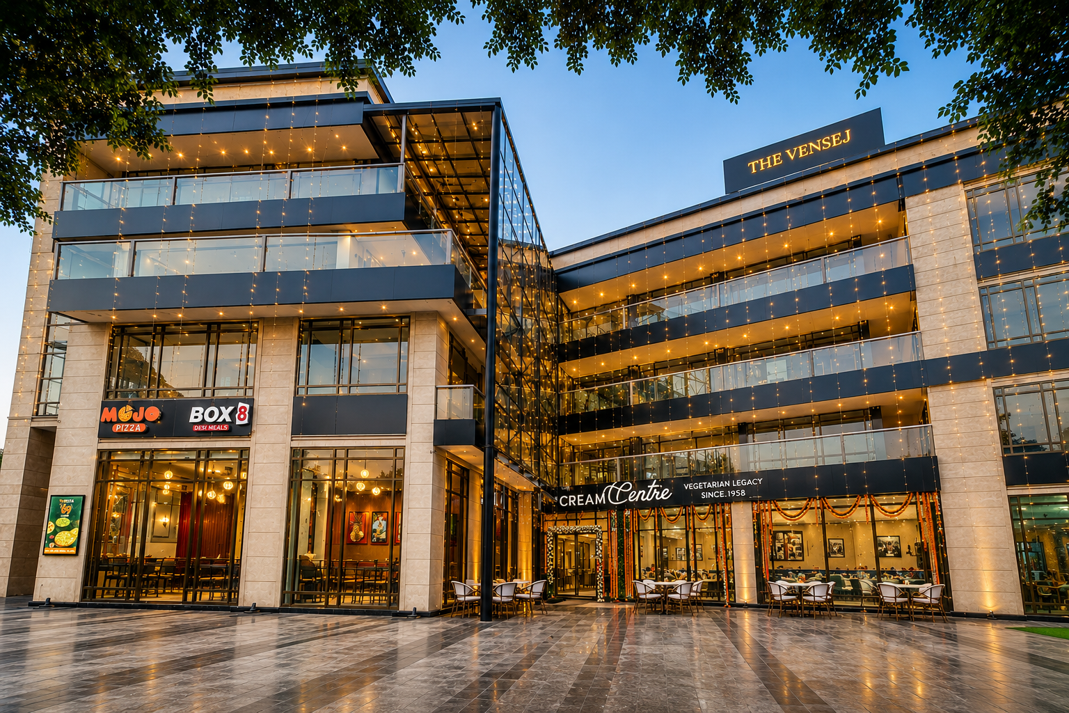 Evening exterior view of The Vensej Mall Gurgaon featuring modern architecture, restaurants, and vibrant lighting on Golf Course Extension Road.