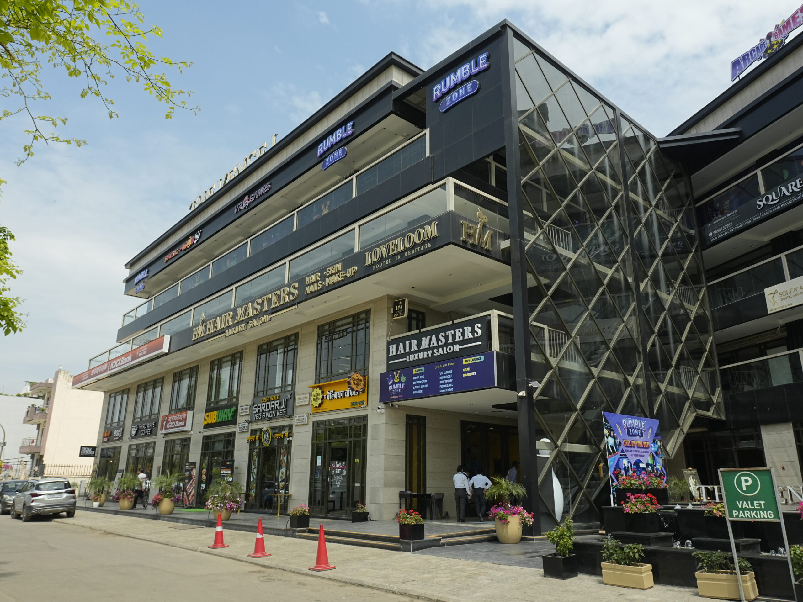 Modern commercial building exterior of The Vensej Mall Gurgaon featuring multiple retail stores, glass architecture, salon signage, gaming zone, and valet parking area under a clear daytime sky.